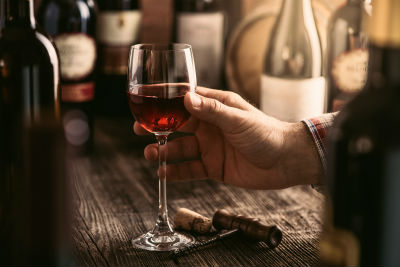 Man holding wine glass on wood table with bottles in background and cork at base of glass